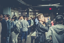 A crowded indoor gathering with people socializing and some holding drinks. The setting appears to be a casual and relaxed environment with dim lighting and a mix of individuals in conversation.