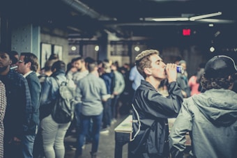 A crowded indoor gathering with people socializing and some holding drinks. The setting appears to be a casual and relaxed environment with dim lighting and a mix of individuals in conversation.