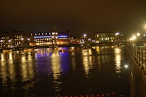 A nighttime cityscape featuring a river with reflections of illuminated buildings and streetlights. The architecture includes buildings with prominent windows and a bridge crossing over the water, displaying vibrant blue and yellow lights. The scene conveys a peaceful urban atmosphere with a softly lit promenade alongside the river.