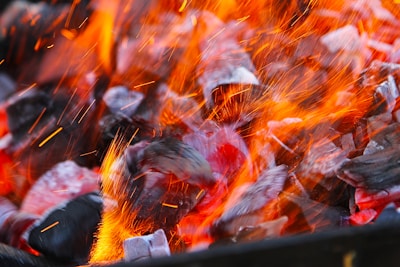 Glowing hot coals with bright orange flames and sparks flying upwards. The coals display a range of red and gray hues, with a dynamic interplay of light and heat.