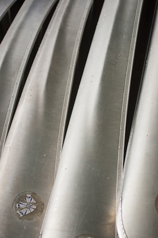 A close-up view of several metallic structures aligned in parallel, with a smooth and shiny surface. There are visible bolts or rivets along the edges of the metallic panels, and the surface reflects light at various angles.