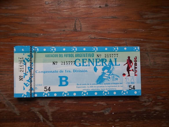 A vintage ticket for an Argentine football match, with blue and white colors. The ticket features text indicating 'Asociaci&oacute;n del Futbol Argentino' along with 'Campeonato de 1ra. Divisi&oacute;n'. There is an image of a football player on the right side. The ticket includes numbering and series information.