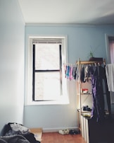 A small, tidy room with light blue walls featuring a single window letting in natural light. Clothes hang neatly on a wooden rack beside a potted plant. A pile of clothes rests on the floor, alongside a pair of shoes. A wooden shelf contains various items like a hat and some boxes.