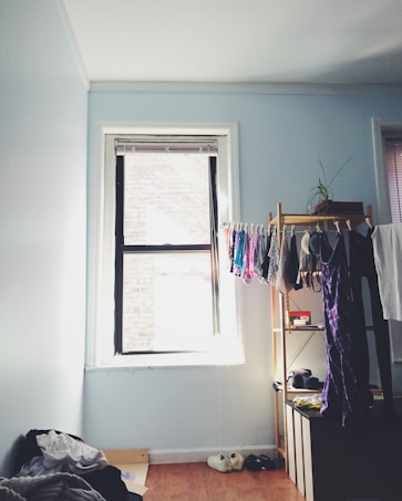 A small, tidy room with light blue walls featuring a single window letting in natural light. Clothes hang neatly on a wooden rack beside a potted plant. A pile of clothes rests on the floor, alongside a pair of shoes. A wooden shelf contains various items like a hat and some boxes.