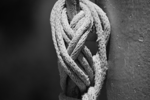 A tightly woven rope with intricate braiding is wrapped around a sturdy pole, showcasing its durability and craftsmanship. The image is in black and white, emphasizing the texture and details of the rope fibers.