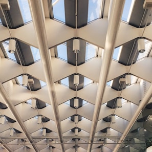 A modern architectural ceiling design with repeated geometric patterns of glass and metal beams. Skylights allow natural light to illuminate the space, while cylindrical lights are evenly distributed across the ceiling.