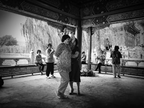 Two people are dancing under a beautifully ornate pavilion with intricate woodwork. In the background, others are standing or seated, possibly watching the dancers or engaged in conversation. The setting seems to be a spacious area near a body of water with trees and traditional architecture visible.