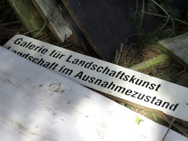 A rustic sign with German text is surrounded by natural elements such as wooden planks, branches, and pine needles. The setting suggests an outdoor or forest environment with uneven surfaces and a mix of light and shadow.