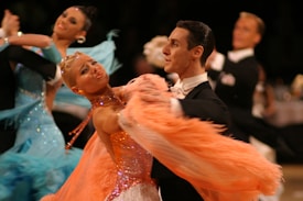 A couple dressed in elegant ballroom attire is captured mid-dance. The woman wears a vibrant, feathered orange and sparkly dress, while the man complements her in a classic black suit and white bow tie. In the background, another couple in similar formal wear, including a striking blue gown with embellishments, can be seen also engaged in the dance.