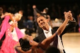 A couple elegantly dressed in formal attire performing a ballroom dance, with the man in a black tuxedo and the woman in a purple gown. They are gracefully holding hands and smiling, surrounded by other dancers in vibrant costumes. The background is blurred, focusing attention on the dancing couple.