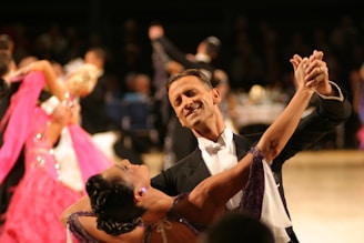 A couple elegantly dressed in formal attire performing a ballroom dance, with the man in a black tuxedo and the woman in a purple gown. They are gracefully holding hands and smiling, surrounded by other dancers in vibrant costumes. The background is blurred, focusing attention on the dancing couple.