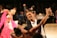 A couple elegantly dressed in formal attire performing a ballroom dance, with the man in a black tuxedo and the woman in a purple gown. They are gracefully holding hands and smiling, surrounded by other dancers in vibrant costumes. The background is blurred, focusing attention on the dancing couple.