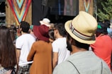 A group of people stands in an outdoor event, likely a concert or festival, as they face a stage with colorful decorations. The audience is diverse, wearing casual summer outfits including hats. The atmosphere appears lively and relaxed.