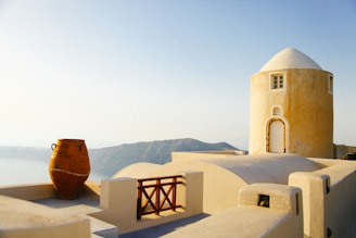 A scenic view featuring a white and ochre cylindrical building with a domed roof situated on a sunlit terrace. A large clay pot decorates the foreground, and there are distant hills under a clear sky in the background.