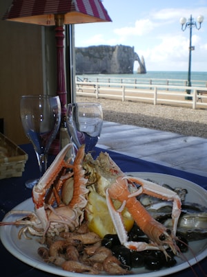 A plate filled with a variety of seafood, including shrimp, prawns, and a large shellfish or crab, is set on a table. There is a slice of lemon in the midst of the seafood. In the background, through the window, a scenic view of the ocean and a cliff can be seen. A lamp with a red and white striped shade is partially visible, and two empty wine glasses are on the table.