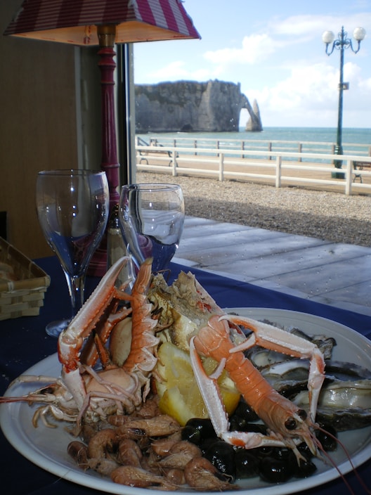 A plate filled with a variety of seafood, including shrimp, prawns, and a large shellfish or crab, is set on a table. There is a slice of lemon in the midst of the seafood. In the background, through the window, a scenic view of the ocean and a cliff can be seen. A lamp with a red and white striped shade is partially visible, and two empty wine glasses are on the table.