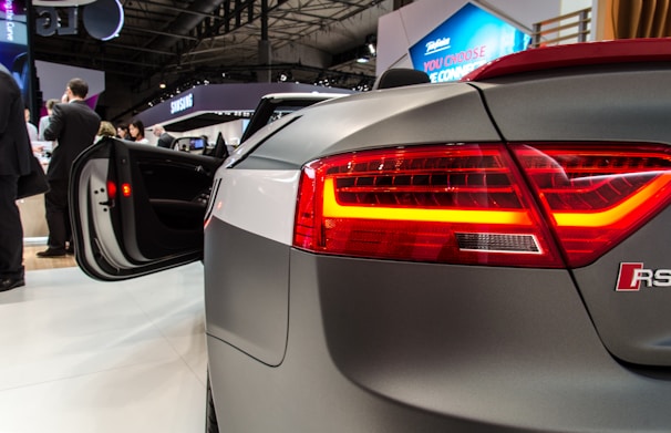 A sleek, modern car with a high-tech design is displayed at an exhibition. The open car door and illuminated taillights catch attention. In the background, several people are engaged in conversation, and there are visible brand logos on banners and signs.