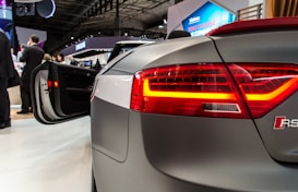 A sleek, modern car with a high-tech design is displayed at an exhibition. The open car door and illuminated taillights catch attention. In the background, several people are engaged in conversation, and there are visible brand logos on banners and signs.