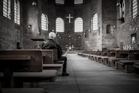 A person with gray hair sits alone in a large, empty church with high ceilings. The interior features exposed brick walls, arched windows allowing natural light, and wooden pews. An altar is visible at the front, adorned with a cross and various church ornaments.