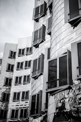 A modern architectural building with a metallic facade, featuring numerous rectangular windows with varied angles. The structure has a wavy, reflective surface that creates interesting light patterns.