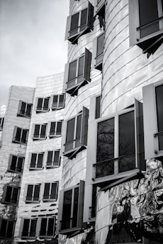 A modern architectural building with a metallic facade, featuring numerous rectangular windows with varied angles. The structure has a wavy, reflective surface that creates interesting light patterns.