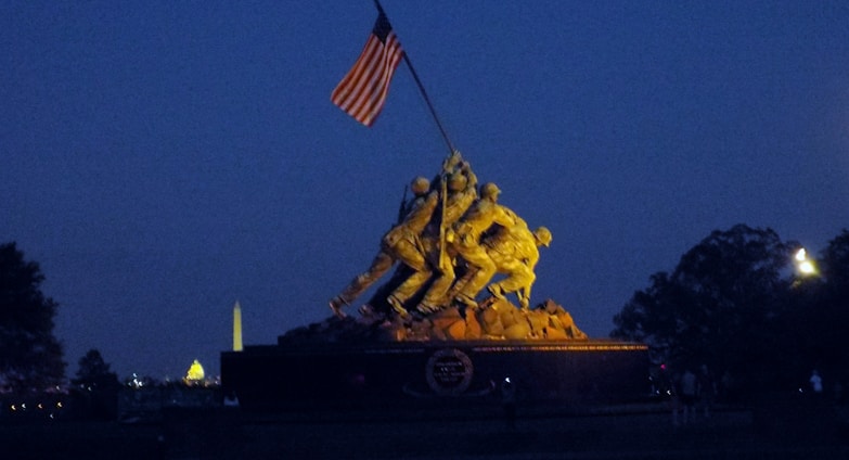 A monument depicting soldiers raising a flag, set against a night sky with city lights visible in the background.