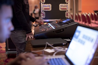 A person is operating a large audio mixing console which has multiple knobs and sliders, combined with a laptop that has a blurred out screen in the foreground. In the background, there are technical racks and some empty red chairs arranged in rows.