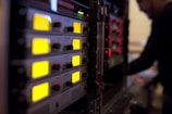 A series of electronic audio devices with illuminated yellow LED screens and small control knobs. The equipment is stacked in a rack format, and a person is visible in the background, slightly blurred, likely operating the gear.