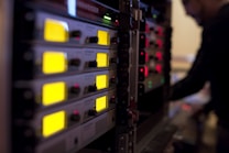 A series of electronic audio devices with illuminated yellow LED screens and small control knobs. The equipment is stacked in a rack format, and a person is visible in the background, slightly blurred, likely operating the gear.