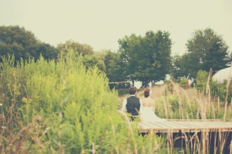 A couple is sitting together on a wooden platform surrounded by lush green vegetation. One person is wearing a white dress while the other is dressed in a black vest and white shirt. In the background, there are several trees creating a serene and natural setting.