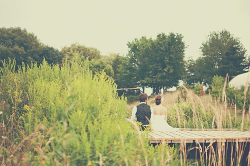 A couple is sitting together on a wooden platform surrounded by lush green vegetation. One person is wearing a white dress while the other is dressed in a black vest and white shirt. In the background, there are several trees creating a serene and natural setting.
