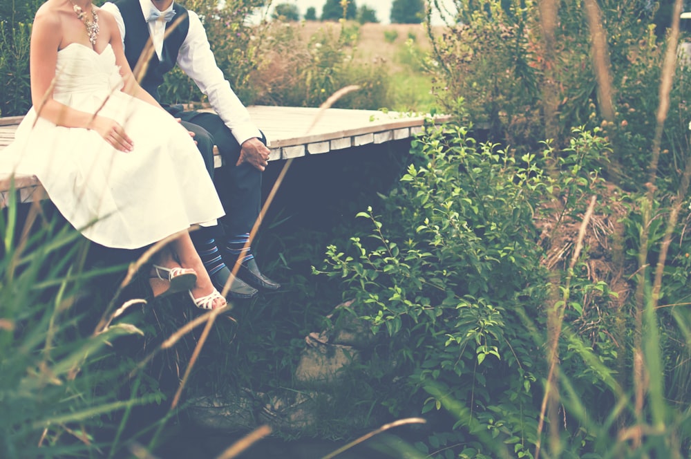 A bridal couple sitting on a wooden footbridge surrounded by green grasses and bushes
