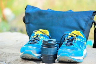 A pair of blue athletic shoes with yellow accents sits on a stone surface next to a camera lens. Behind them, a blue fabric bag is partially visible, suggesting an outdoor or travel setting.