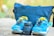 A pair of blue athletic shoes with yellow accents sits on a stone surface next to a camera lens. Behind them, a blue fabric bag is partially visible, suggesting an outdoor or travel setting.