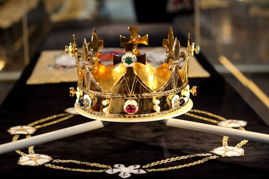 A lavishly decorated golden crown adorned with various gemstones and ornate designs sits prominently on a black surface. The crown features fleurs-de-lis and crosses, with colorful jewels embedded around its circumference. There is an intricate pattern beneath the crown, resembling a ceremonial cloth with gold and white embroidery.