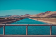 A serene aqueduct flowing through a desert landscape with a clear blue sky above. Mountains and dry hills form the backdrop, while the surrounding area features sparse vegetation and agricultural lands.
