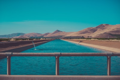 A serene aqueduct flowing through a desert landscape with a clear blue sky above. Mountains and dry hills form the backdrop, while the surrounding area features sparse vegetation and agricultural lands.
