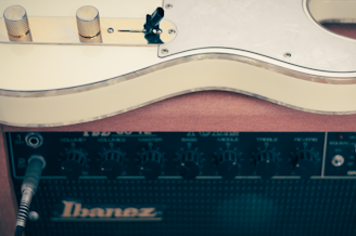 A cream-colored electric guitar rests on top of a black Ibanez amplifier. The guitar is viewed partially, focusing on its body and control knobs. The amplifier features various control dials for volume, bass, and treble settings.