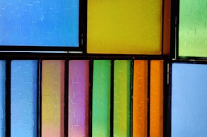 A composition of stained glass panels divided by dark frames, featuring vibrant colors such as blue, green, orange, yellow, and pink. The glass has a textured, frosted appearance creating a mosaic effect.