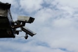 Two security cameras mounted on the corner of a building against a partly cloudy sky. The cameras are positioned at different angles, likely for surveillance purposes.