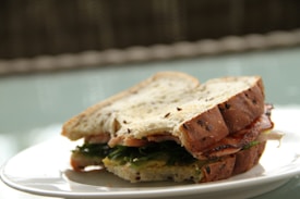 A sandwich made with multigrain bread is placed on a white plate. The sandwich is filled with layers of fresh lettuce and visible slices of meat.