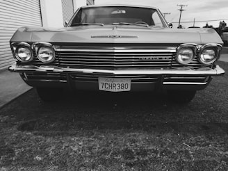 A classic car with a vintage design and prominent round headlights is parked on a textured concrete surface. The front grille and bumper display shiny, chrome details. A California license plate is visible, adding to the retro aesthetic.