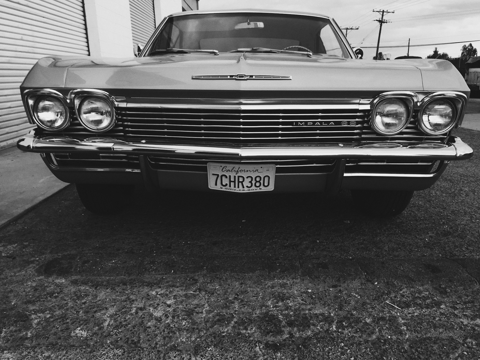A classic car with a vintage design and prominent round headlights is parked on a textured concrete surface. The front grille and bumper display shiny, chrome details. A California license plate is visible, adding to the retro aesthetic.