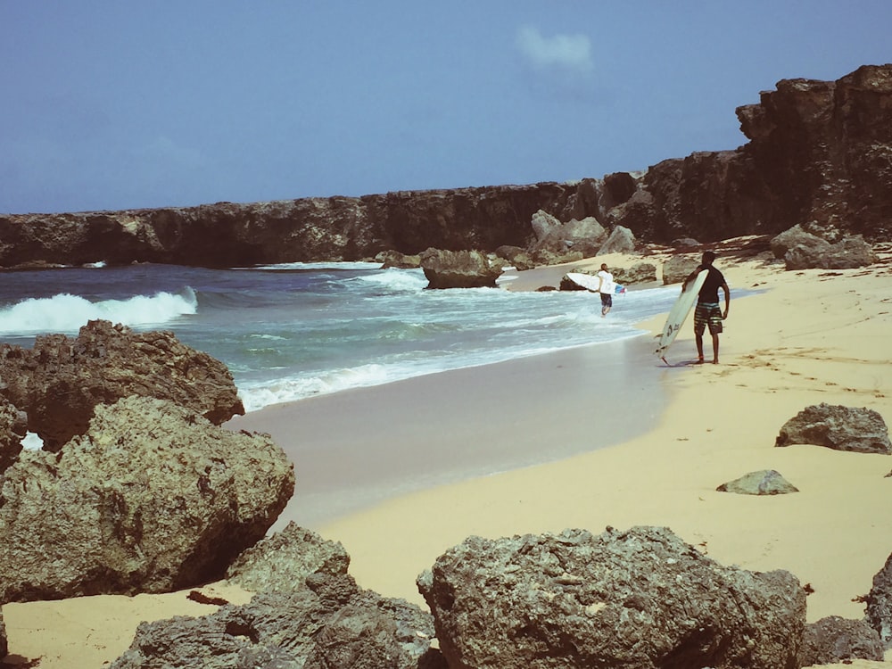 person wearing black top and gray shorts standing on shore holding surfboard