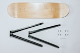 A wooden skateboard deck is positioned horizontally at the top, with a simple logo on one end. Below it, two black metal brackets are visible, designed for assembling a furniture piece, likely a table or shelf. Six screws are neatly arranged in two rows on the right, accompanied by a small Allen wrench.