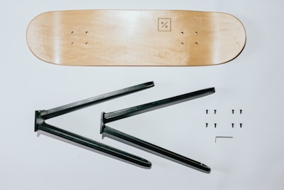 A wooden skateboard deck is positioned horizontally at the top, with a simple logo on one end. Below it, two black metal brackets are visible, designed for assembling a furniture piece, likely a table or shelf. Six screws are neatly arranged in two rows on the right, accompanied by a small Allen wrench.