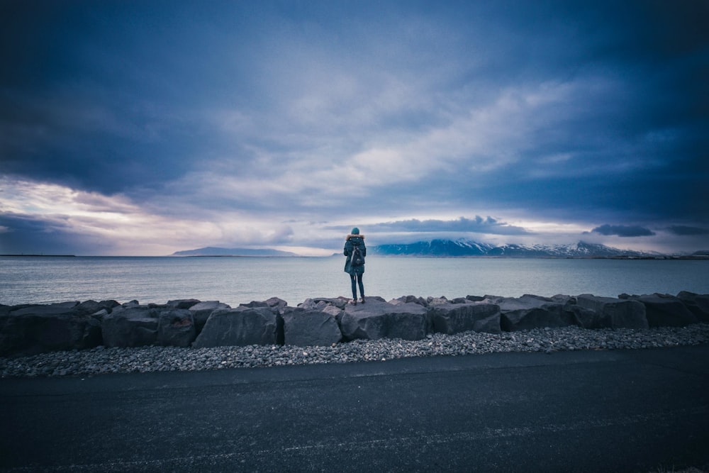 photo of person standing on rock looking sea during daytime