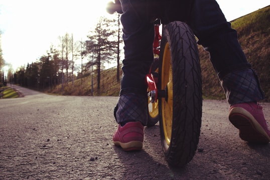 A child on a bicycle is riding along a paved path. The focus is on the rear wheel of the bike, which is prominently in the foreground. The path is surrounded by trees and grassy areas, with sunlight filtering through, creating a serene outdoor setting.