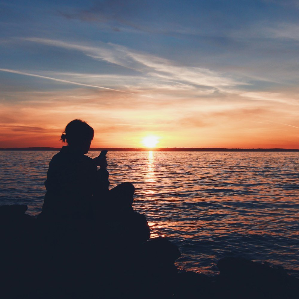 silhouette photo of person sitting on boulder near body of water