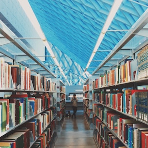 A modern library interior featuring long aisles lined with bookshelves filled with various colorful books. The scene is framed by a high, geometric ceiling with a striking blue pattern. In the center of the aisle, a person is taking a photograph, adding a touch of dynamic presence to the serene environment.